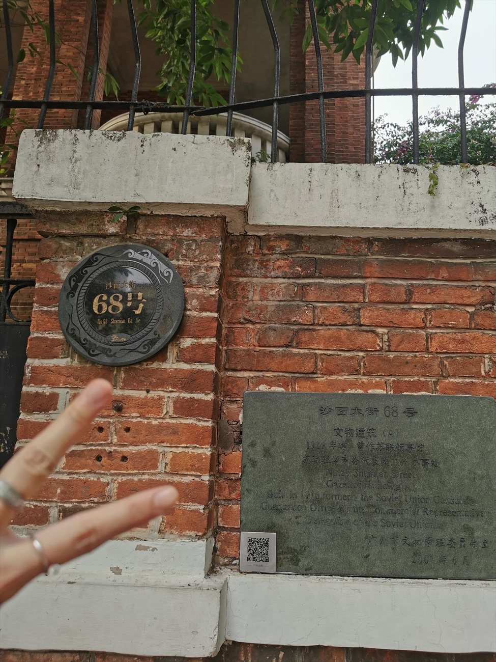 2024年，一只手在沙面大街68号的圆形门牌与铭牌前比出拍照手势